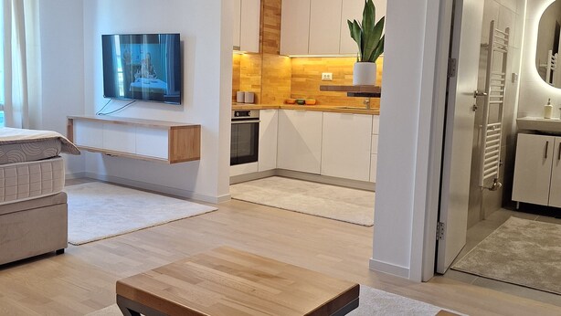 A modern living room with a sofa, a coffee table, and a TV. A kitchen is visible in the background with wooden cabinets and a countertop. - Sunset Dream Apartment (Sarajevo)