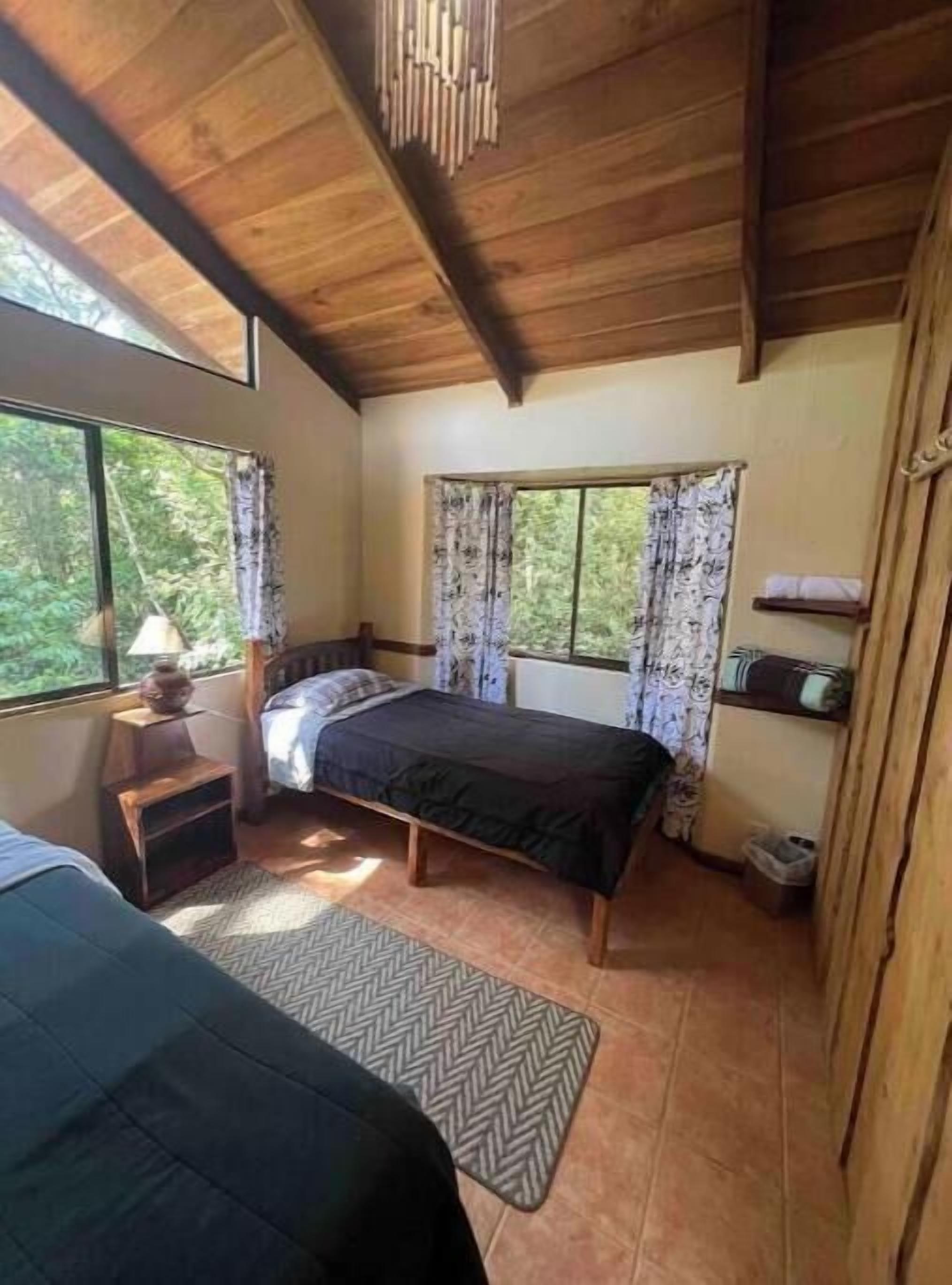 Una camera da letto con un letto, un comodino, una finestra con tende e vista sul verde.