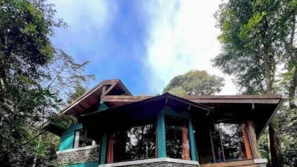 A two-story house with a stone facade and a wooden balcony, surrounded by lush greenery.