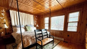 A wooden cabin bedroom with a four-poster bed, a chair, and a window with blinds.