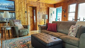 A cozy living room with wooden walls, a brown leather sofa, a matching ottoman, a patterned rug, and a painting of birds on the wall.