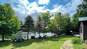 A lakeside cabin with a stone pathway leading to a picnic table and chairs, surrounded by trees and a view of the lake.