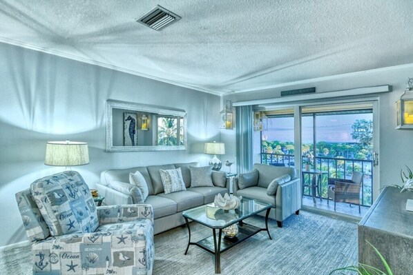 A modern living room with a grey sofa, armchairs, and a glass coffee table. There is a large window with a view of the outdoors.