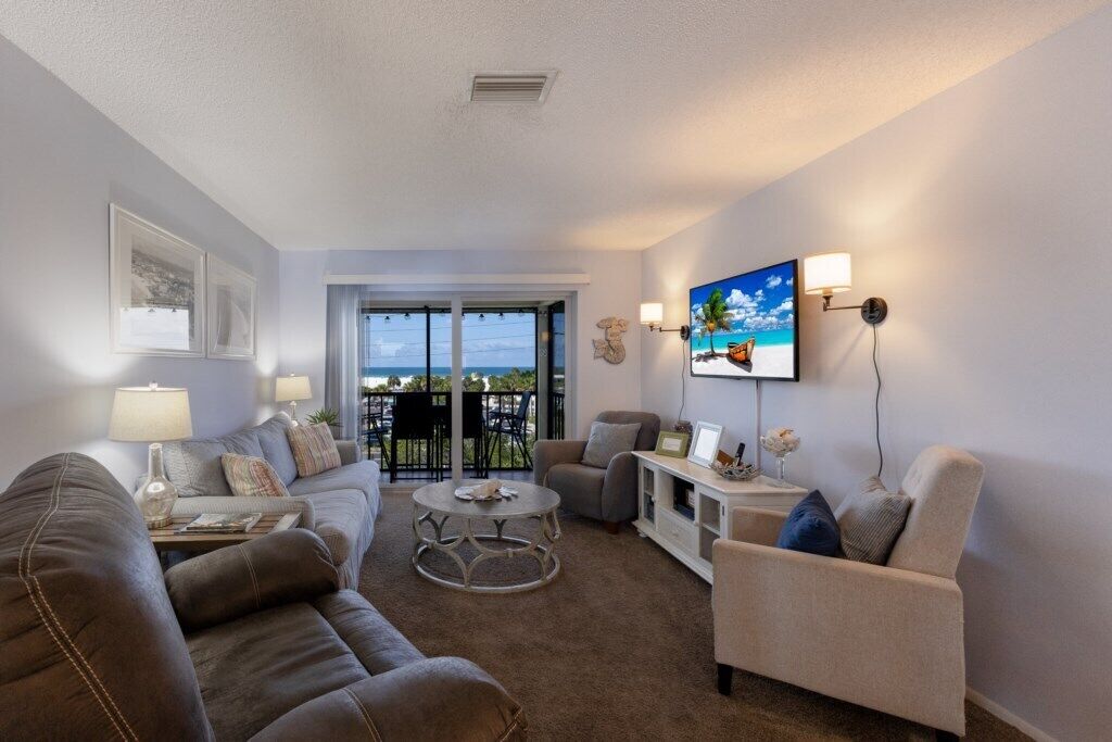Ein Wohnzimmer mit einer Couch, Sesseln, einem Couchtisch und einem an der Wand befestigten Fernseher. Es gibt einen Balkon mit Blick auf den Ozean.