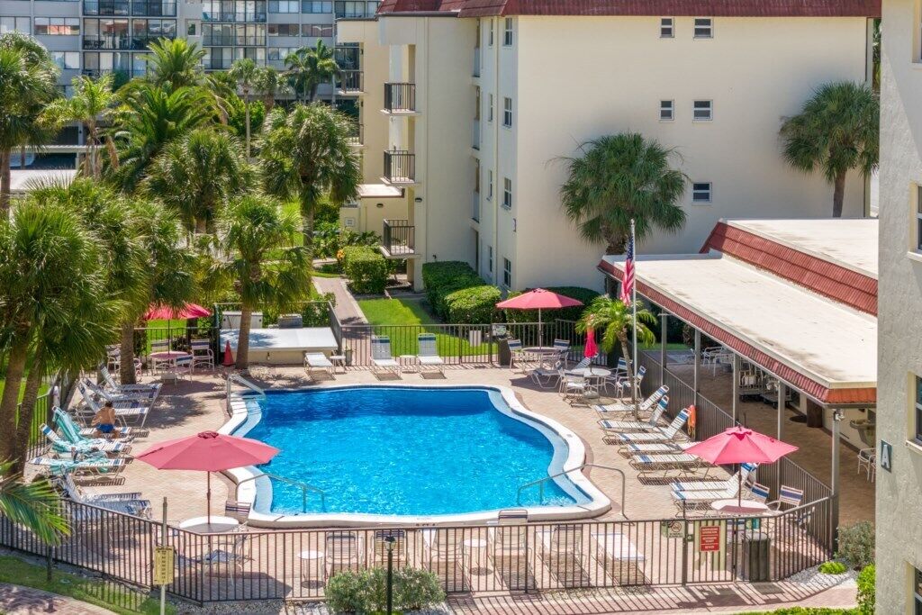 Ein Swimmingpool, umgeben von Liegestühlen und Sonnenschirmen, mit Palmen und einem Gebäude im Hintergrund.