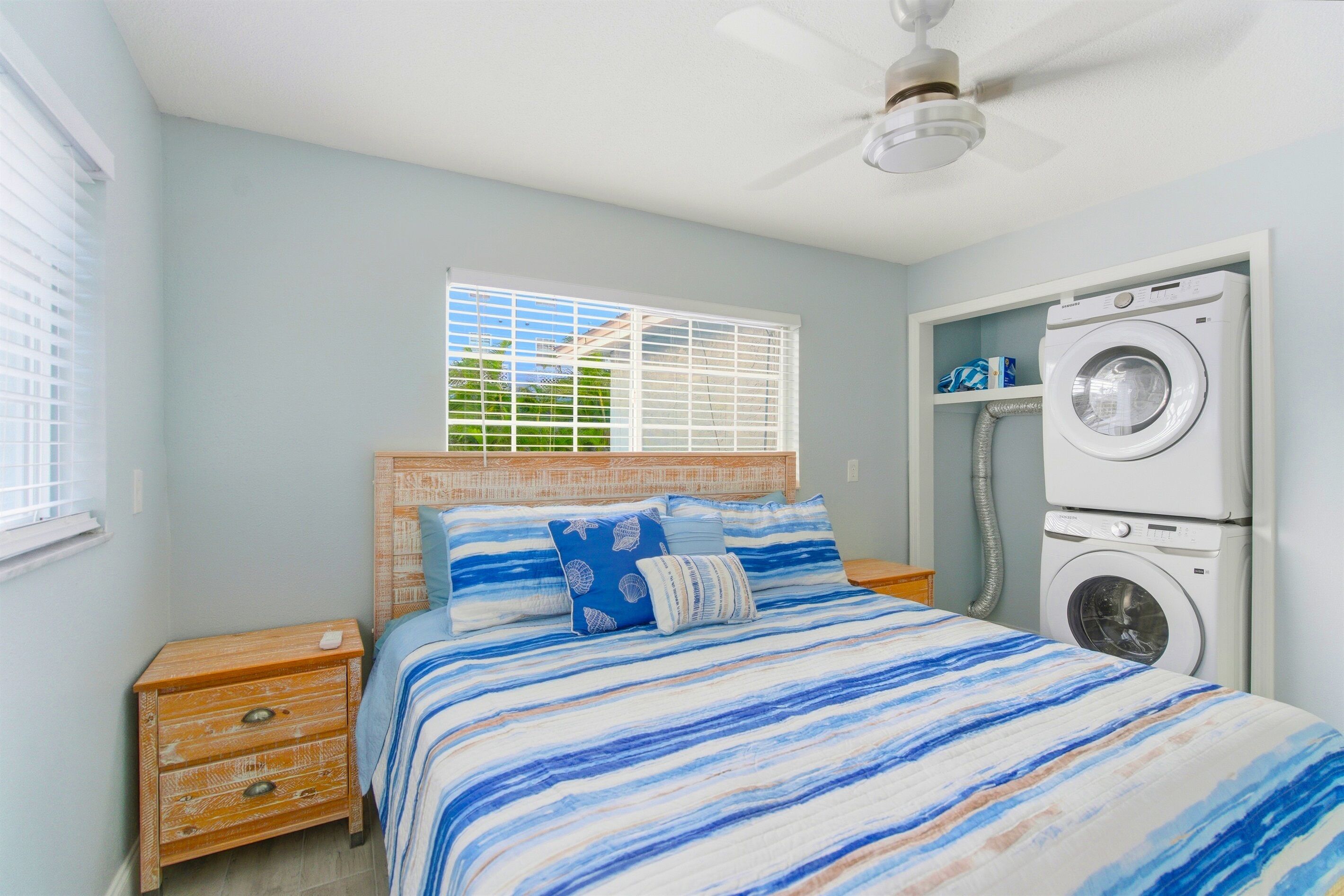 A bedroom with a bed, a nightstand, a ceiling fan, a window with blinds, and a built-in washer and dryer.