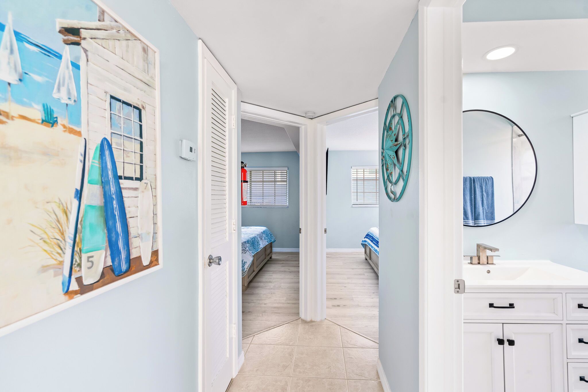 Un pasillo con un mural alusivo a la playa, una puerta blanca, una cama con colcha azul y blanca a rayas, y un baño con un espejo redondo y un lavabo.