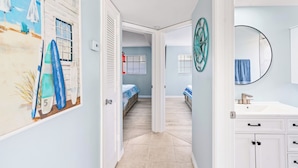 Un couloir orné d’une fresque murale sur le thème de la plage, une porte blanche, un lit recouvert d’une couvre-lit à rayures bleues et blanches, et une salle de bain avec un miroir rond et un lavabo.