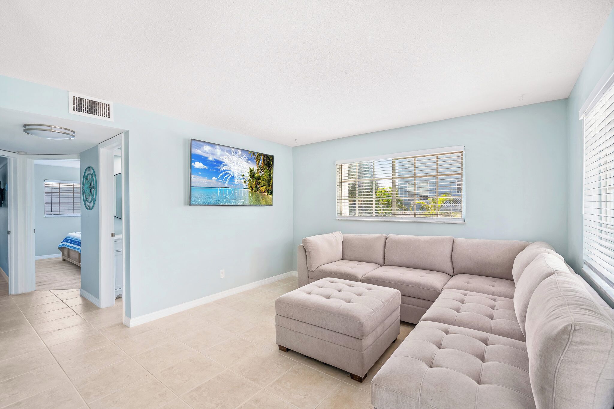 A living room with a sectional sofa, an ottoman, a large window with blinds, and a painting of a beach scene.