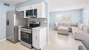 Une cuisine moderne équipée d’appareils électroménagers en acier inoxydable, d’un four à micro-ondes et d’une cuisinière. Un canapé d’angle beige se trouve dans l’espace salon, près d’une fenêtre.