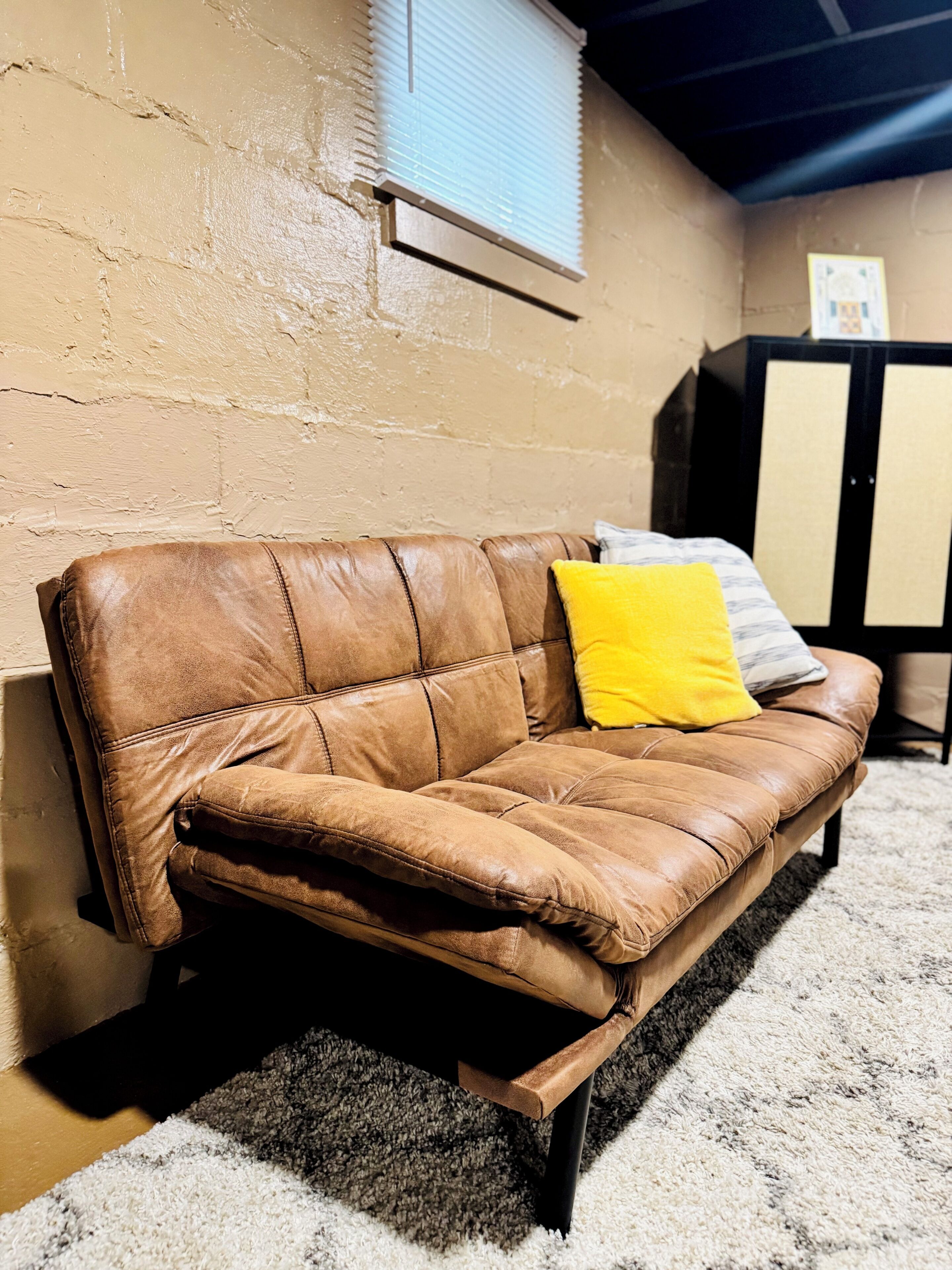 A brown leather sofa with a yellow pillow against a beige wall, a window with blinds, and a dark-colored cabinet in a room with a carpeted floor.