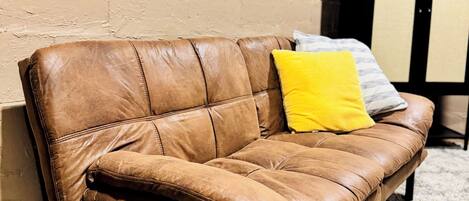 A brown leather sofa with a yellow pillow against a beige wall, a window with blinds, and a dark-colored cabinet in a room with a carpeted floor.