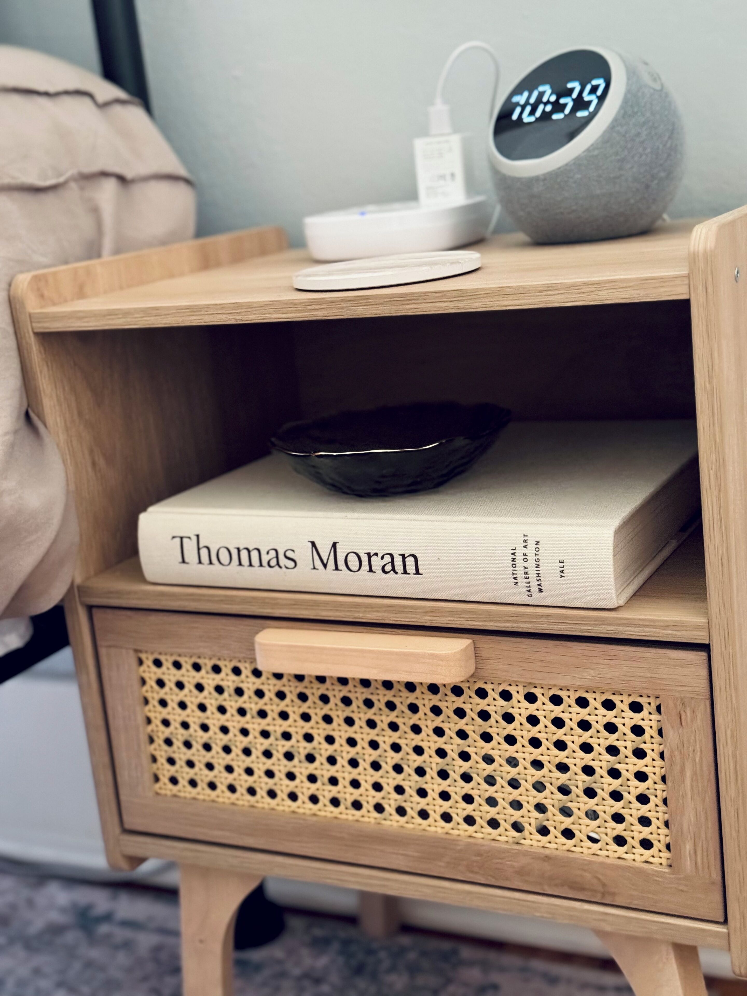 A wooden bedside table with a book titled 'Thomas Moran' and a digital clock displaying 10:39.