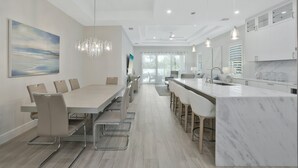 A modern kitchen with a marble island, a dining area with a long table and chairs, and a large painting on the wall. - New! Sunset Walk – Brand New Luxury Home, Just a Short Stroll to Vanderbilt Beach (Naples)