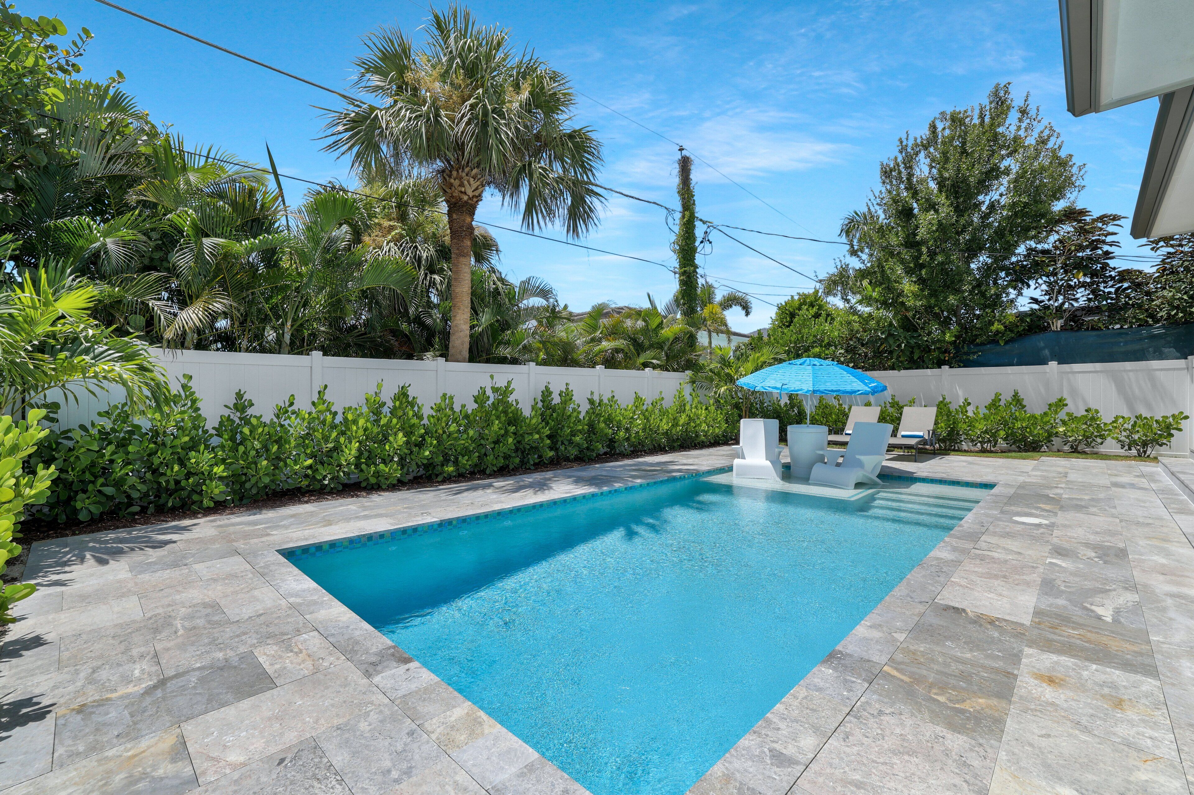 A rectangular swimming pool with a white deck, surrounded by lush greenery and a white fence.
