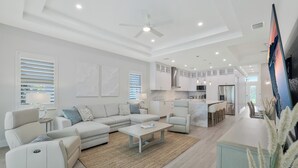 A modern living room with a sofa, a recliner, and a coffee table. There is a kitchen area with cabinets and a dining table. - New! Sunset Walk – Brand New Luxury Home, Just a Short Stroll to Vanderbilt Beach (Naples)