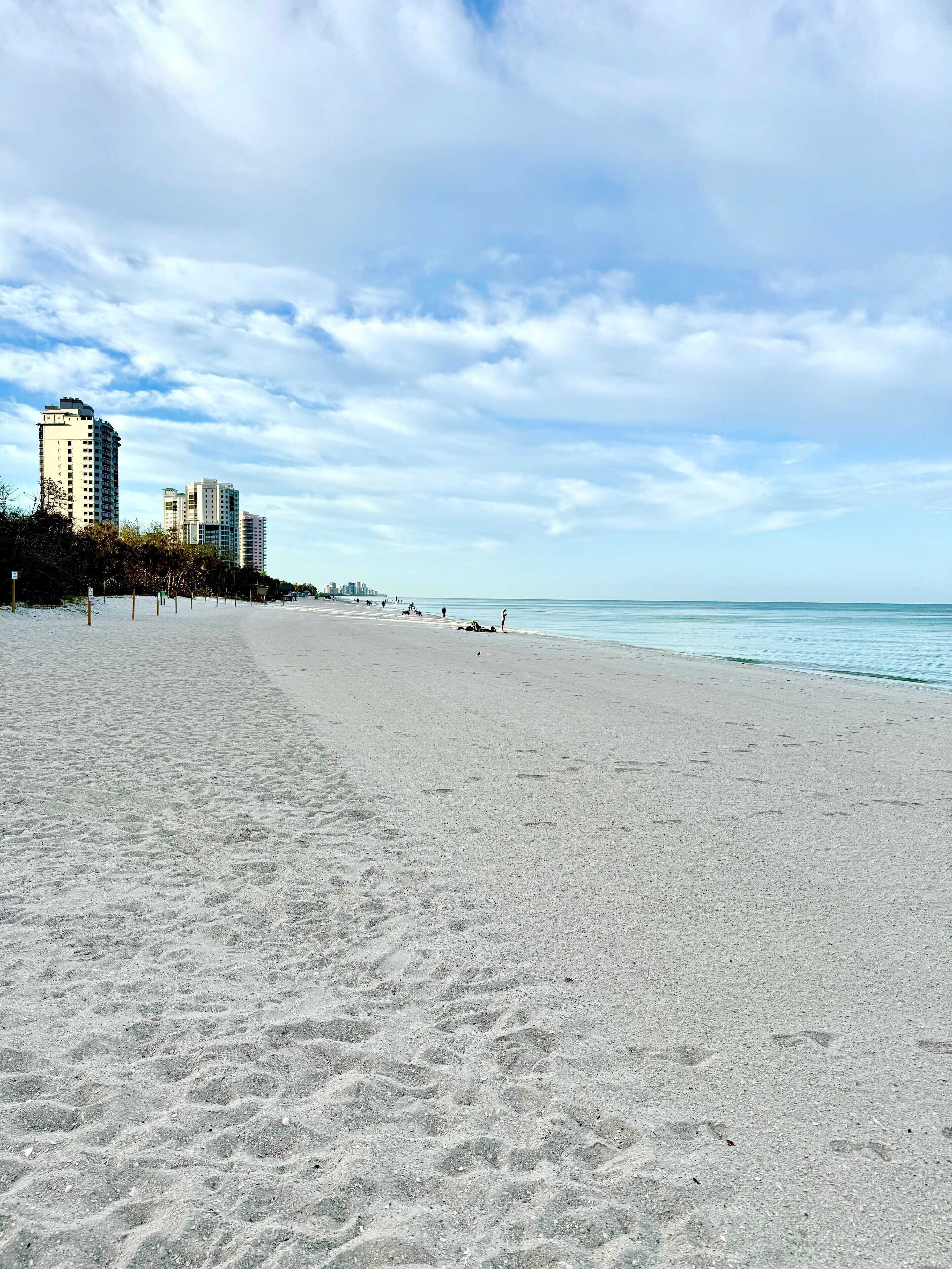 A wide sandy beach with footprints, a clear sky, and buildings in the background.