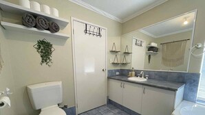 Ein Badezimmer mit Toilette, Waschbecken und Dusche. Handtücher und ein Spiegel sind auf Regalen zu sehen.