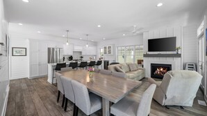A modern kitchen with white cabinetry, a large island with seating, and a dining area with a wooden table and chairs. - NEW Lakeside Cottage Sleeps 16 Includes Golf Cart Walk to Lake and Near Pool (Lakeside)