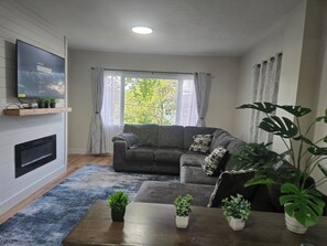 Uma sala de estar com sofá cinza, televisão de tela plana na parede, mesa de centro de madeira com plantas em vasos e uma grande janela com cortinas.
