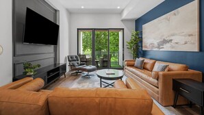 A modern living room with a large TV, leather sofas, a coffee table, and a view of the outdoors through large sliding doors. - PFR Suites - Butler|2-Story Lawrenceville Penthouse w/ Rooftop Terrace|2 King Beds /2.5 Bath|Parking (Pittsburgh)