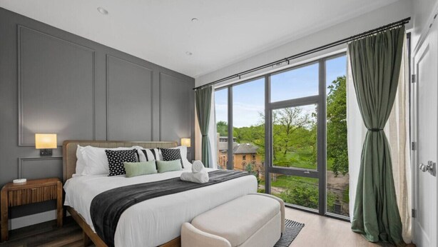 Un dormitorio moderno con una cama grande, una mesita de noche, un banco y vistas a zonas verdes a través de amplios ventanales.