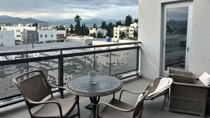 Ein Balkon mit Korbmöbeln und einem Glastisch mit Blick auf eine Stadtlandschaft aus Gebäuden und Bergen.