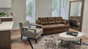 Ein modernes Wohnzimmer mit Sofa, Sessel und Couchtisch. Große Fenster mit Jalousien, eine Küchenzeile und ein Spiegel an der Wand sind ebenfalls vorhanden.