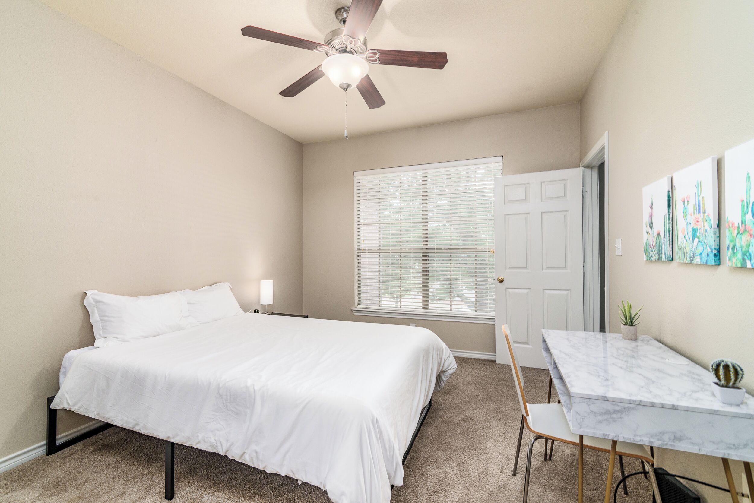 Une chambre avec un lit, un ventilateur de plafond, une fenêtre avec des stores, une porte, une petite table avec une chaise et un cactus.
