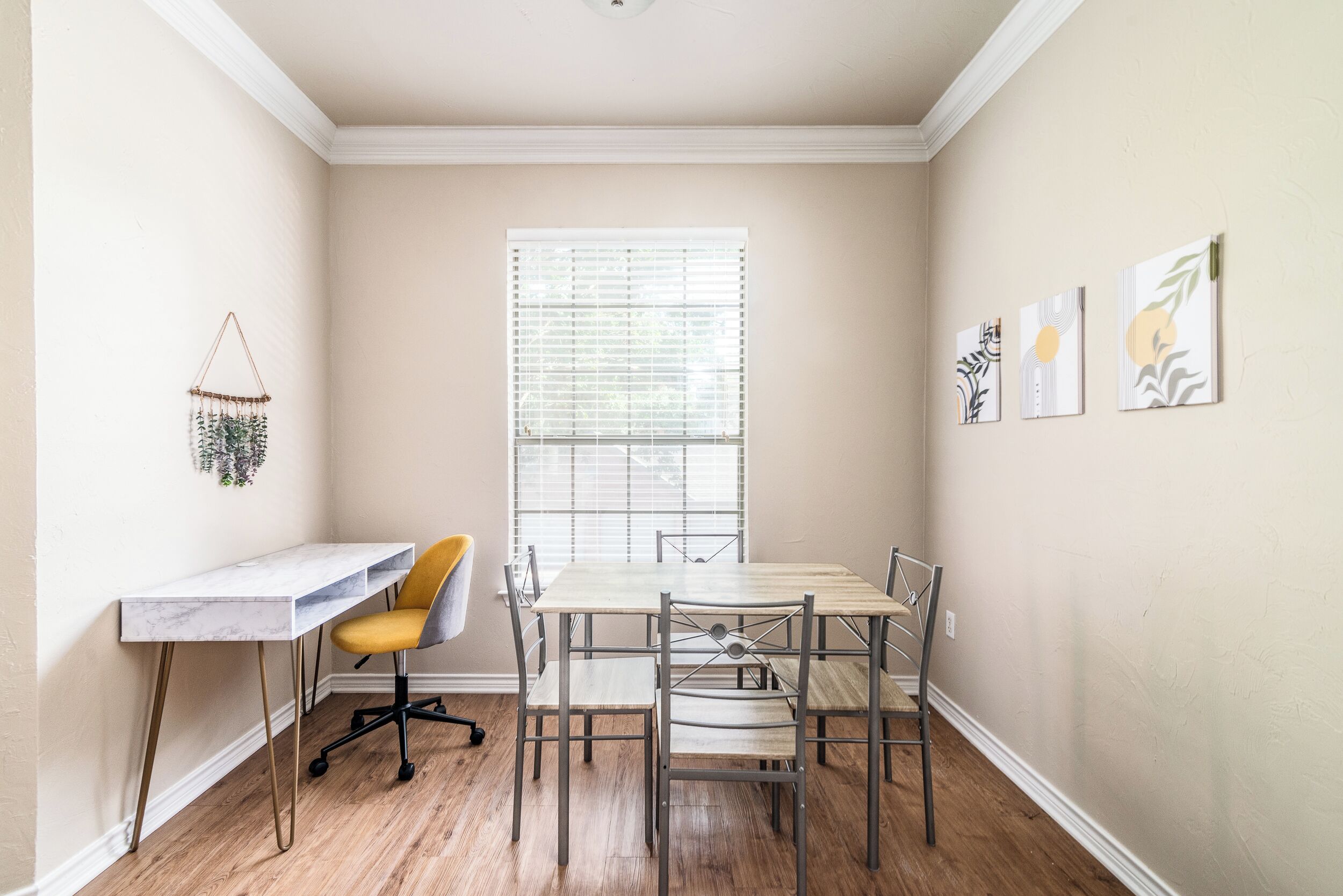 A dining area with a table and chairs, a small desk with a chair, a window with blinds, and wall art.