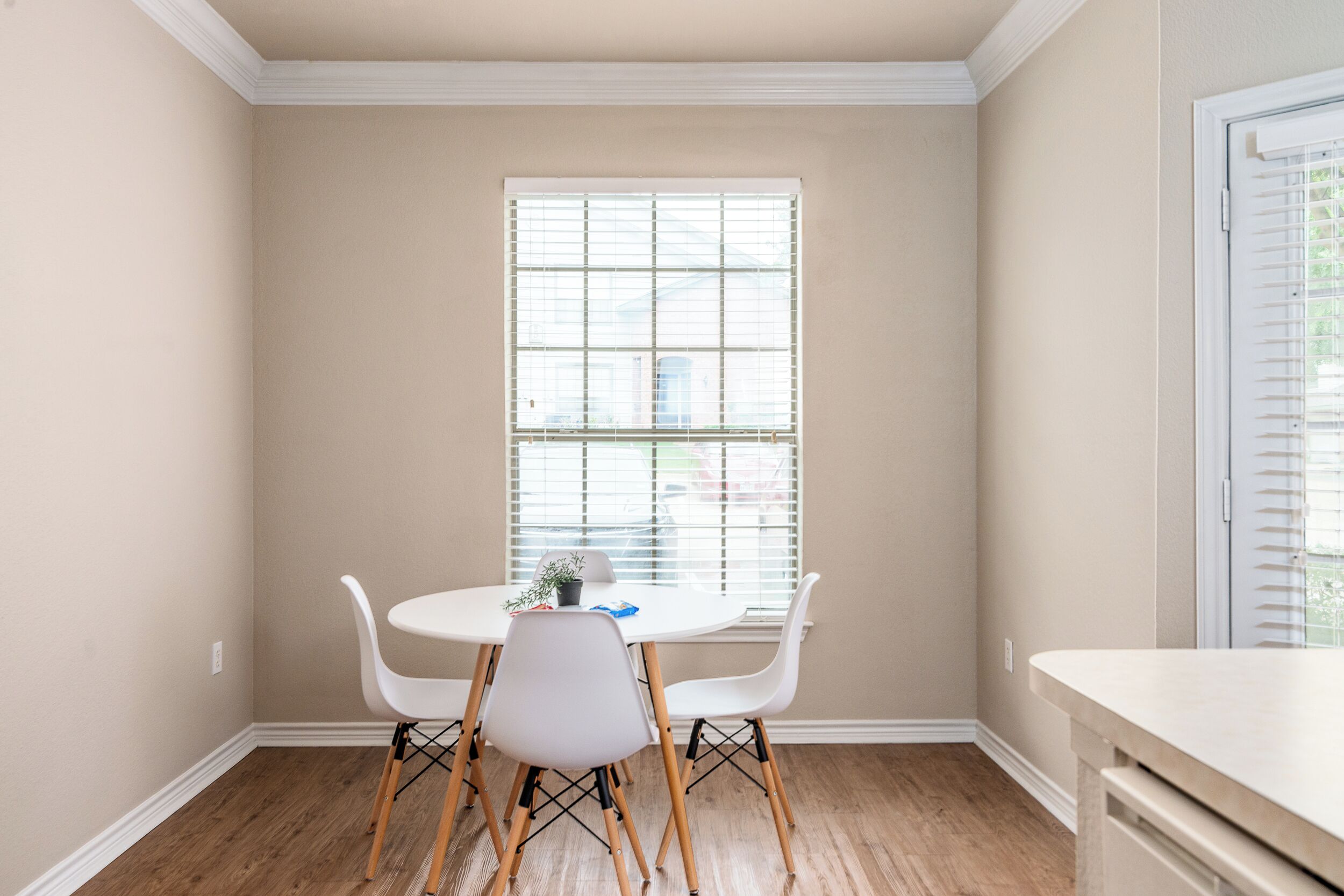 A dining area with a round table, two white chairs, and a window with blinds.
