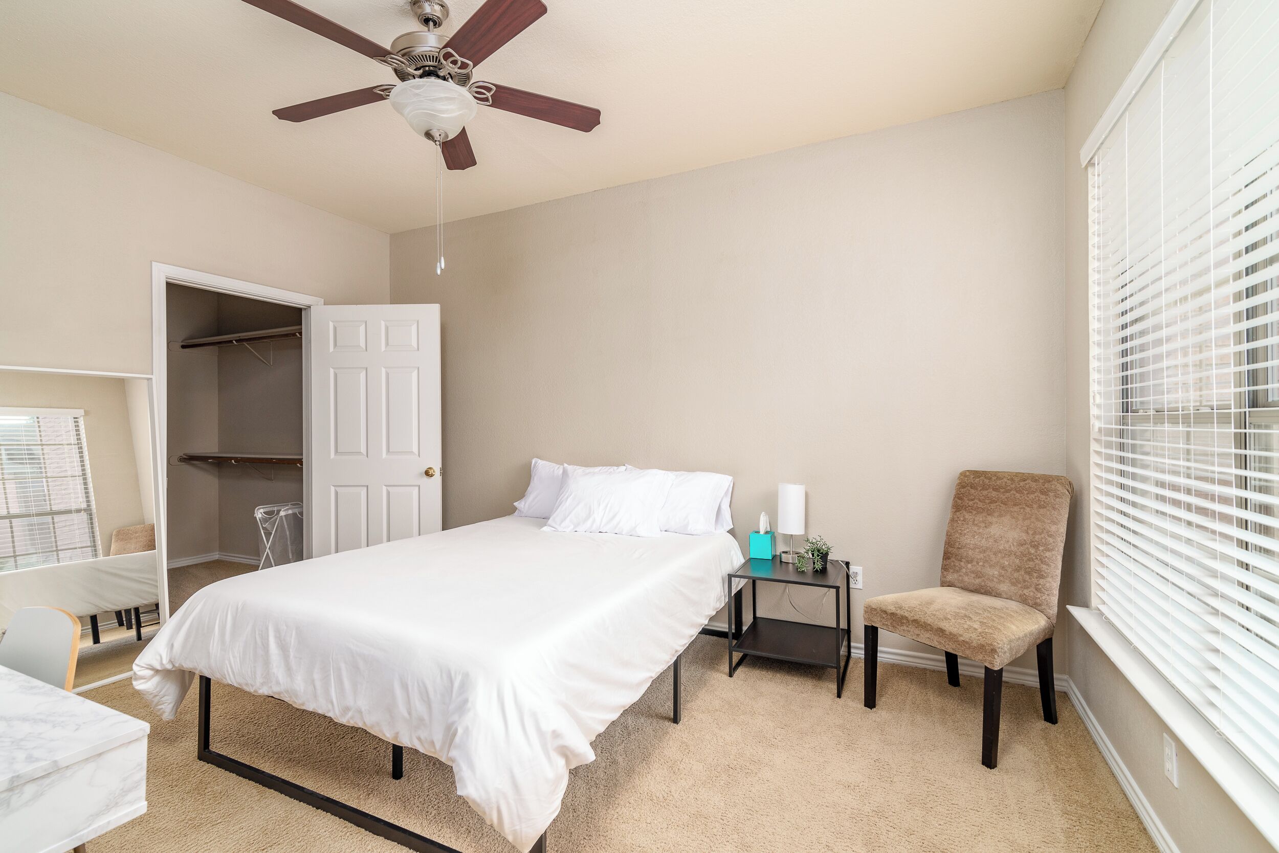 A bedroom with a bed, a chair, a nightstand, a ceiling fan, a closet, and a window with blinds.
