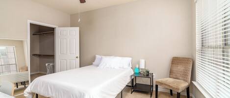 A bedroom with a bed, a chair, a nightstand, a ceiling fan, a closet, and a window with blinds.