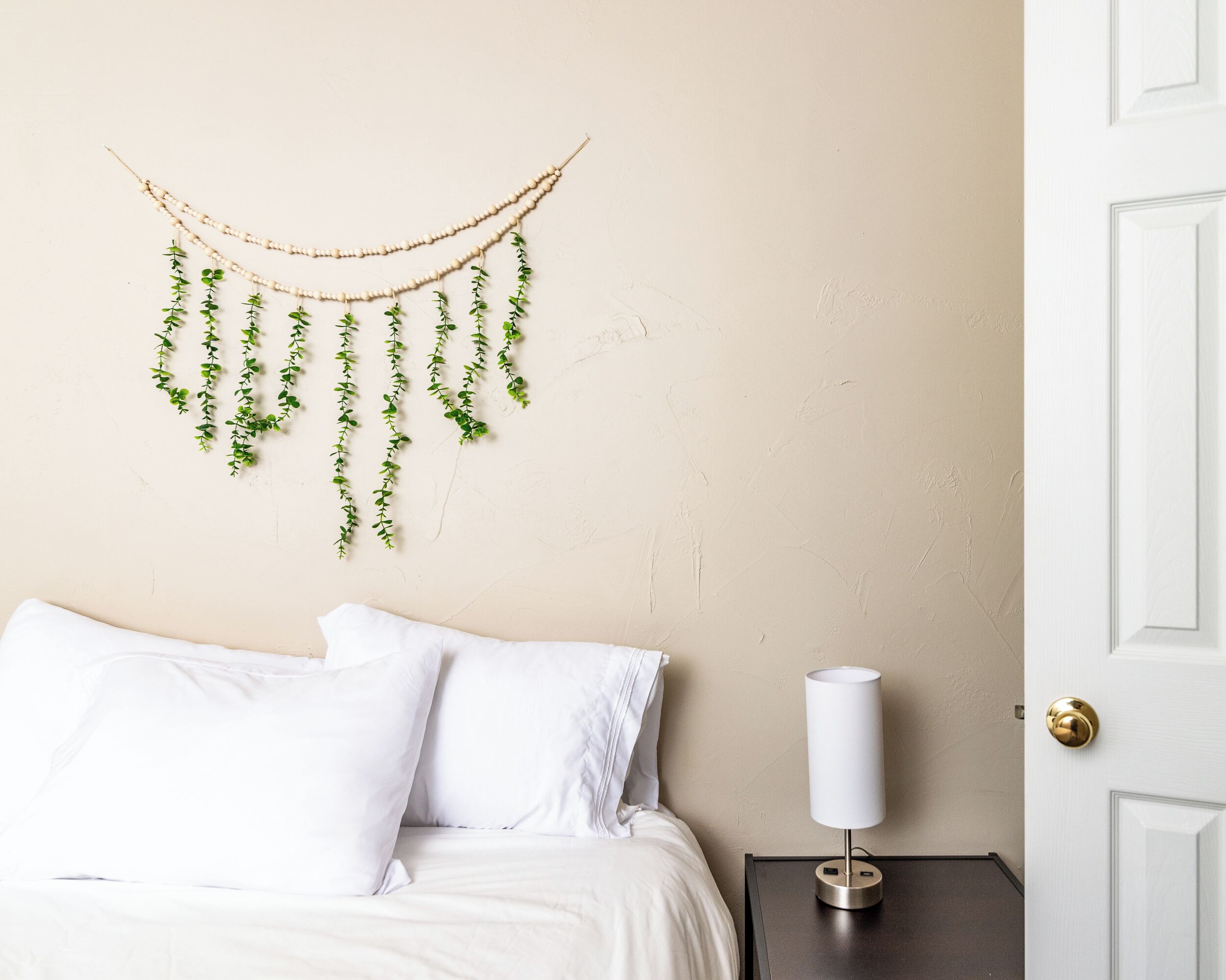 A bedroom with a bed, white pillows, a bedside table with a lamp, and a wall-mounted plant decoration.