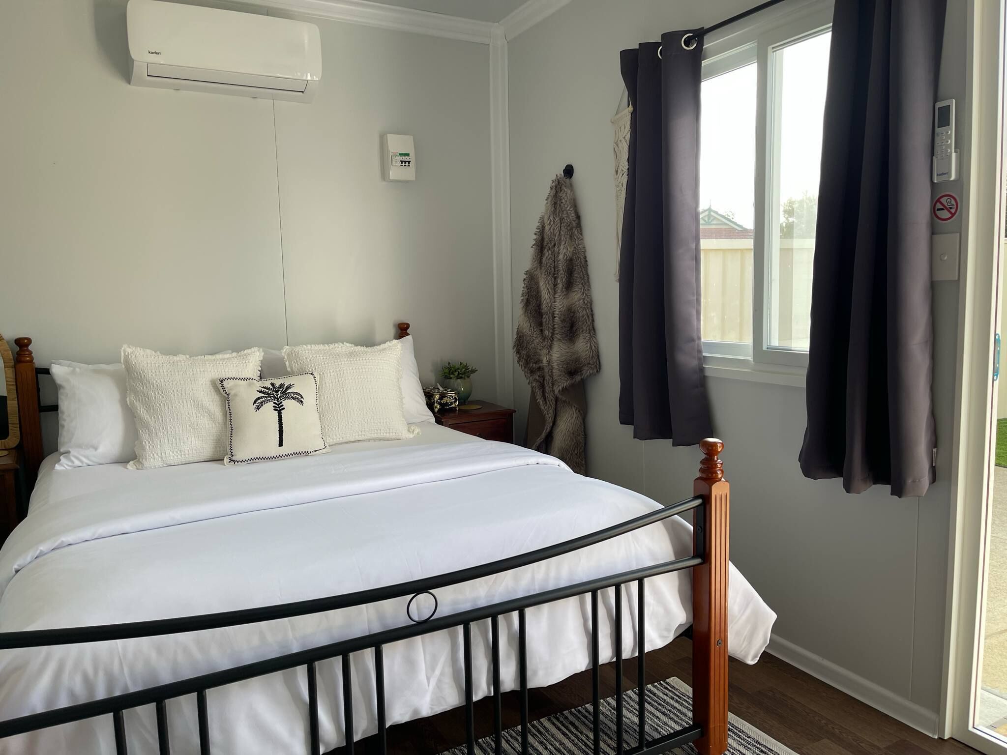 Un lit bien fait, avec une literie blanche et un coussin orné d’un motif de palmier, une fenêtre avec des rideaux foncés et un climatiseur fixé au mur.