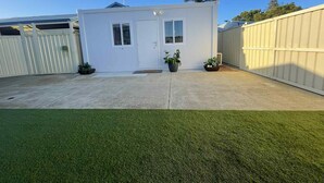 A small, single-story building with a white exterior, a front door, and two windows. There are two potted plants on either side of the door. The building is surrounded by a white fence and a green lawn. - Wannanup Retreat Short Stay Accommodation. Pet-Friendly! (Wannanup)