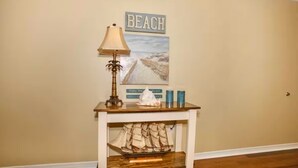 A room with a wooden table, a lamp, a ship model, and a beach-themed poster on the wall.