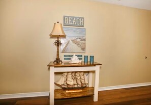 A room with a wooden table, a lamp, a ship model, and a beach-themed poster on the wall.