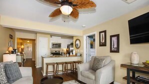 A living room with a ceiling fan, a sofa, a coffee table, and a kitchen area in the background.