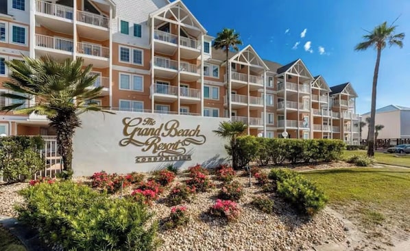 A multi-story hotel with balconies, a sign reading 'The Grand Beach Resort,' and landscaped gardens with shrubs and flowers. - Festival Sale! Amazing Balcony View~Pool & Hot Tub (Gulf Shores)
