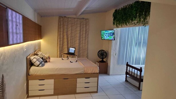 A bedroom with a bed, a laptop on the bed, a TV on the wall, a fan, and a window with curtains. - Vilars154- Knights and Lagoon (Macaé)