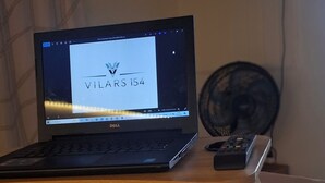 Ein Laptop auf einem Rollgestell, dessen Bildschirm 'VILARS i54' anzeigt. Im Hintergrund befinden sich ein Bildschirm und ein Ventilator.