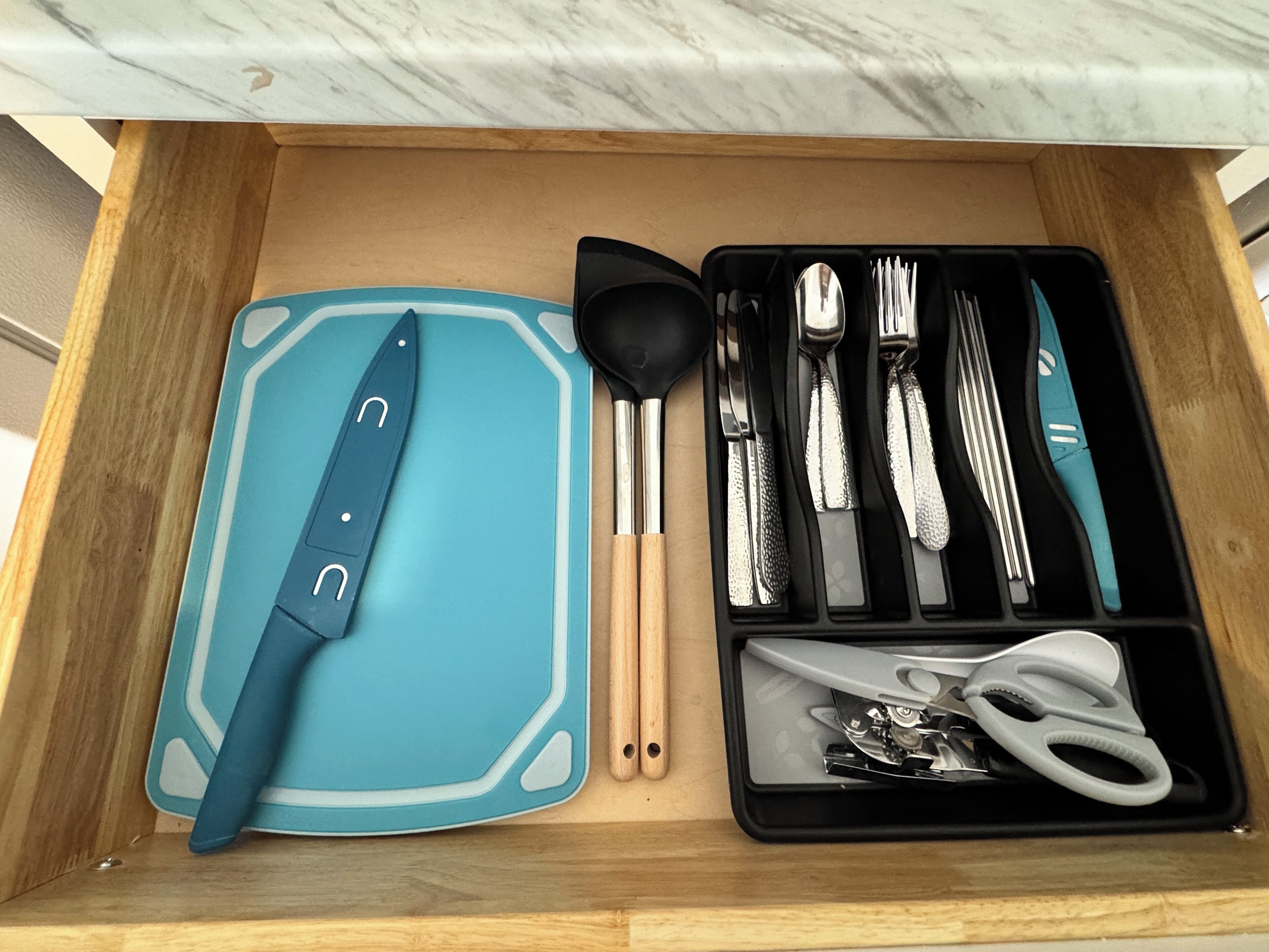 A drawer containing a blue cutting board, a knife, a spoon, a spatula, a set of forks, a set of knives, and a pair of scissors.