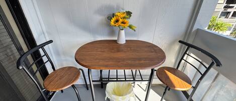 A small balcony with a wooden table and two chairs, a vase with sunflowers, and a white trash bag on the floor.