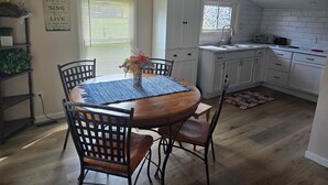 A dining area with a round wooden table, four chairs, and a blue rug. There is a window with blinds, a kitchen with white cabinets, and a decorative sign on the wall. - Quaint Country Home with Hot Tub (Crown Point)