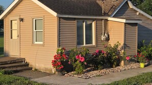 A small, single-story house with a beige exterior, a front porch with steps, and a garden with pink flowers. - Quaint Country Home with Hot Tub (Crown Point)