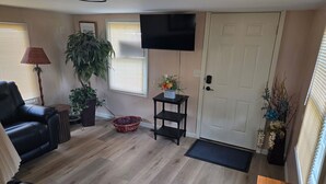 A living room with a black leather sofa, a wooden coffee table, a TV mounted on the wall, a potted plant, and a door with a doormat. - Quaint Country Home with Hot Tub (Crown Point)