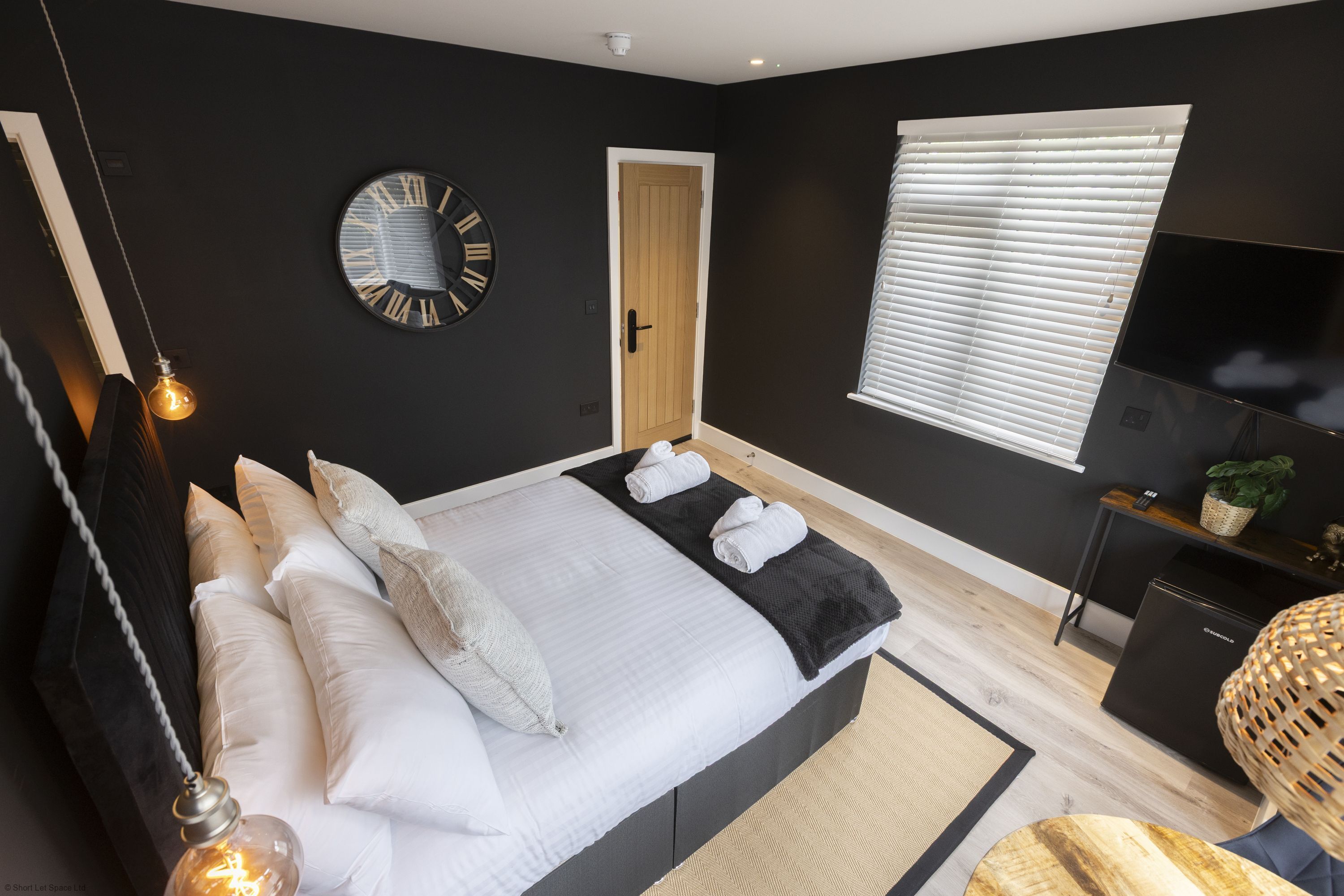 A modern bedroom with a bed, pillows, a round wall clock, a door, a window with blinds, a TV, and a small plant.