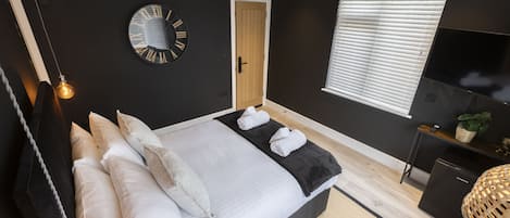 A modern bedroom with a bed, pillows, a round wall clock, a door, a window with blinds, a TV, and a small plant.