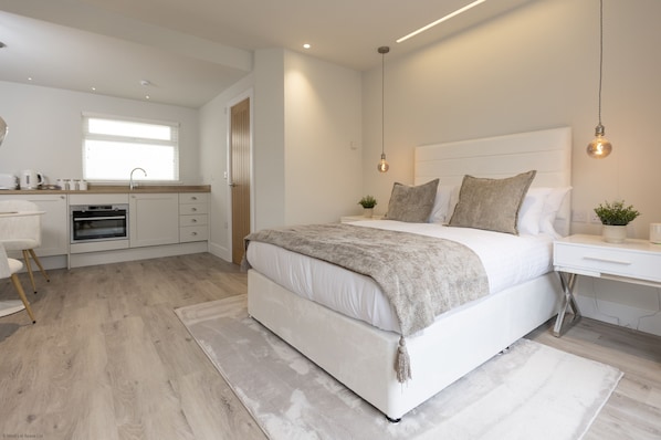 A modern bedroom with a large bed, bedside tables, a desk, and a kitchen area.